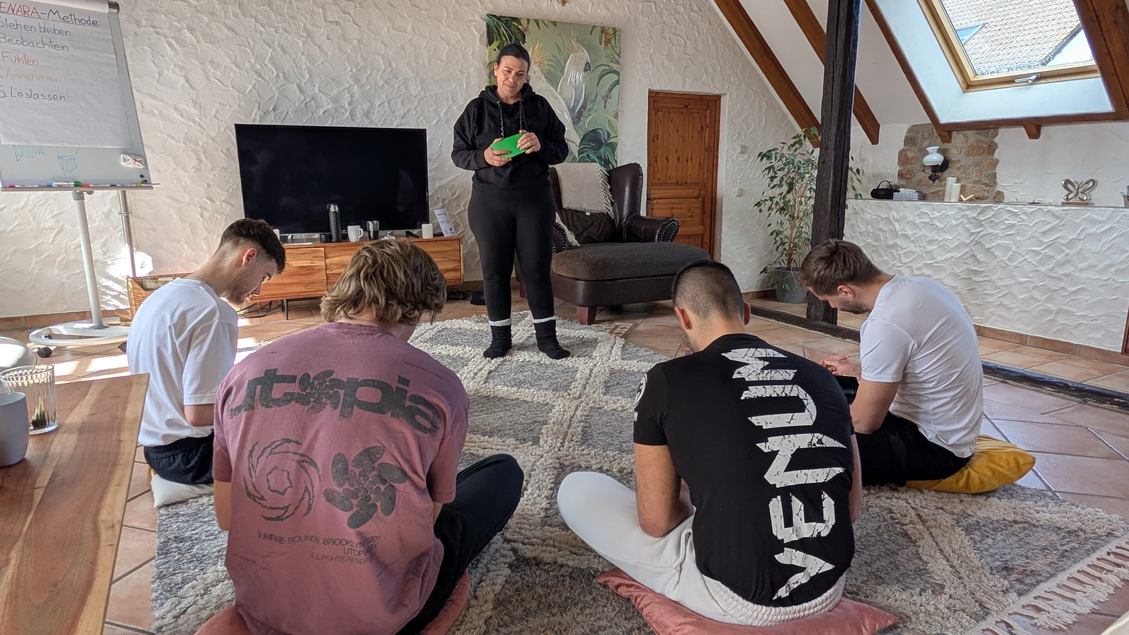 Rosella leitet die Gruppe an, die im Kreis auf dem Boden sitzt; am Flipchart stehen die fünf Schritte der SENARA-Methode